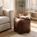 A dark brown square leather ottoman utilized as a small coffee table, holding a ceramic mug and an open book in a bright living room.