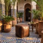 A dark brown square leather ottoman styled outdoors in a sunlit Moroccan courtyard alongside terracotta planters and a brass tea table.