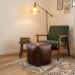 A dark brown square leather ottoman serving as a footrest next to a mid-century green armchair in a cozy reading nook.