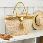 Gold straw bag on white bench, handwoven palm leaf tote with braided gold trim and tassel
