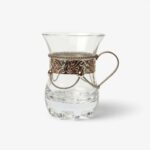 Intricate silver-tone filigree scrollwork on clear glass tea cups with handle showing the pierced metal sleeve