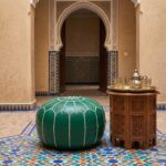 Traditional Moroccan courtyard interior with a green leather floor ottoman next to a carved wooden side table and a tea service.