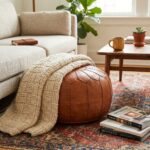 A cozy living room scene with a tan leather round ottoman pouf tucked against a light beige sofa and a chunky knit throw rug.