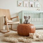 A neutral-toned nursery featuring a round ottoman pouf placed on a fluffy sheepskin rug next to a light wood rocking chair.