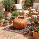A sunny outdoor patio with a round ottoman pouf centered on a blue patterned rug, surrounded by terracotta potted succulents and plants.