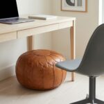 Minimalist home office setup with a round ottoman pouf used as under-desk seating beneath a light wood desk and laptop.
