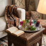 A silver moroccan tea set arranged on a hand-carved wooden side table next to an open book and a cozy Berber-style living room setup.