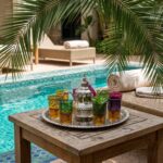Luxury poolside relaxation with a silver moroccan tea set served on a wooden table, overlooking a turquoise tiled swimming pool.