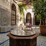 A handcrafted silver tea gift set on a carved wooden table in a sun-drenched courtyard with zellige tilework and orange trees.