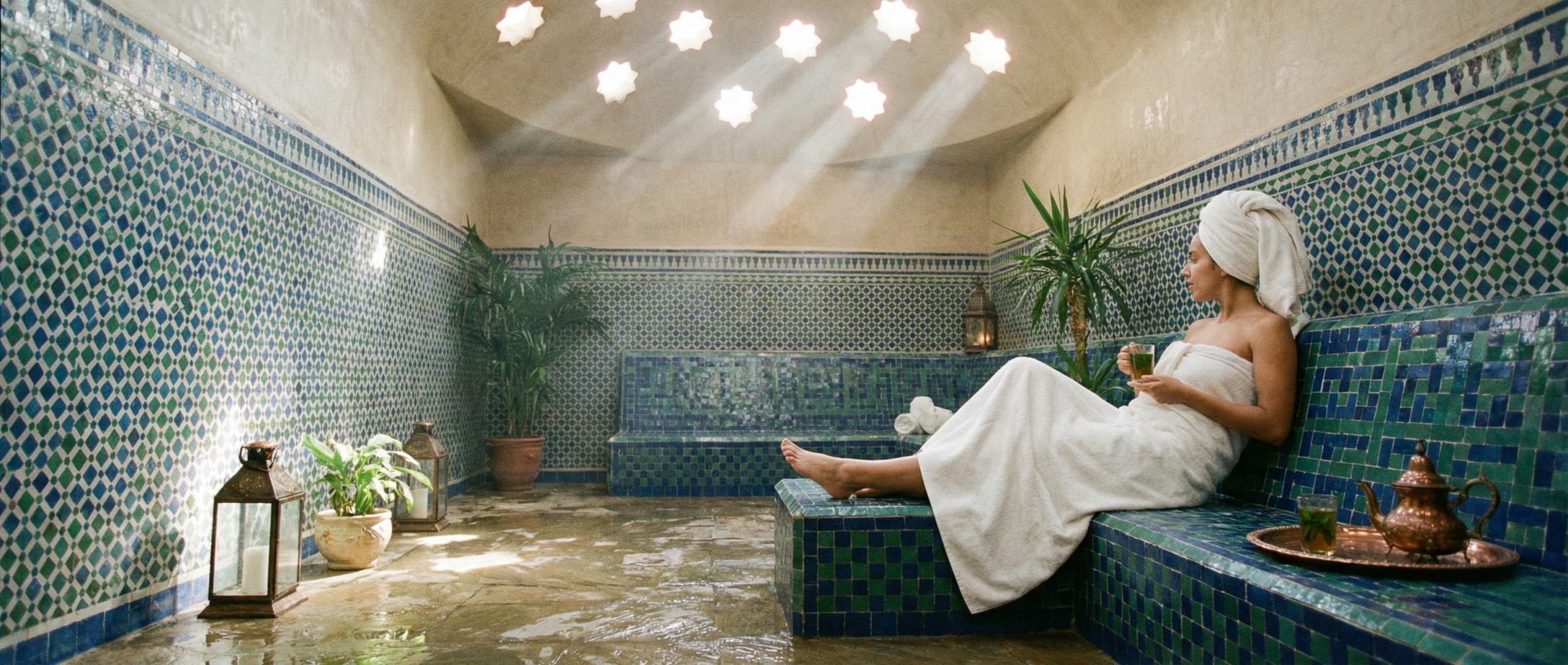 How To Use the Moroccan Hammam A tranquil photo of a person wrapped in a towel, sipping mint tea and resting on a tiled bench in the relaxation area of a traditional Moroccan hammam, with sunlight filtering through a domed ceiling.