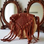 Round boho straw bag with leather fringe displayed on rustic wooden bench
