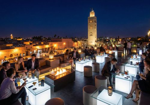 A view from a trendy, crowded rooftop bar at night in Marrakech, looking out over the illuminated ancient city and the Koutoubia Mosque minaret.