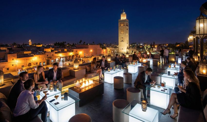 A view from a trendy, crowded rooftop bar at night in Marrakech, looking out over the illuminated ancient city and the Koutoubia Mosque minaret.