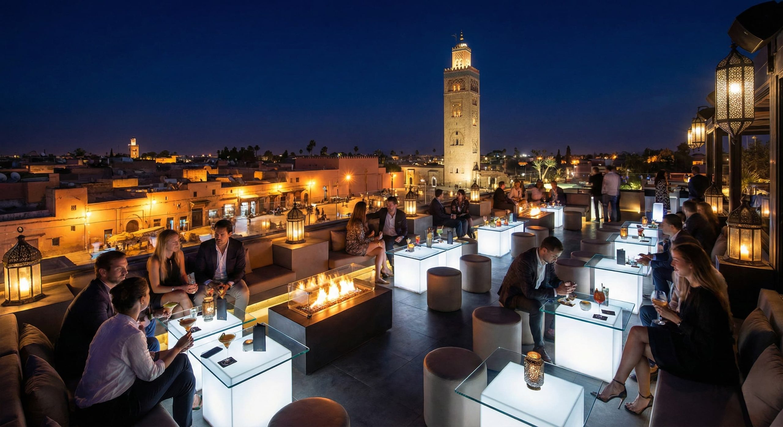 A view from a trendy, crowded rooftop bar at night in Marrakech, looking out over the illuminated ancient city and the Koutoubia Mosque minaret.