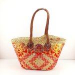 Embroidered straw tote bag with red and yellow floral design, brown leather handles on white background