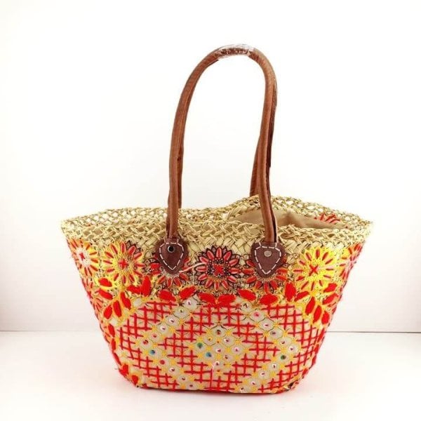 Embroidered straw tote bag with red and yellow floral design, brown leather handles on white background