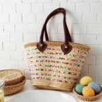 Colorful straw bag beside pool towels showcasing multicolored sequin stripe rows on handwoven palm leaf tote body