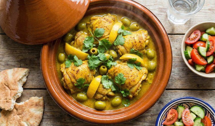 An overhead photograph of a steaming Moroccan chicken tagine in a ceramic pot, filled with chicken, green olives, and preserved lemons in a rich sauce, served with fresh bread on a wooden table.