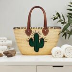 Handwoven palm leaf summer straw bag with embroidered cactus displayed beside white towels and greenery