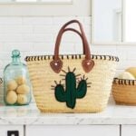 Handwoven palm leaf summer straw bag with cactus motif on marble counter beside glass jar and basket