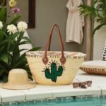 Handwoven palm leaf summer straw bag with green cactus embroidery beside straw hat and sunglasses poolside