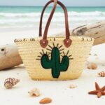 Handwoven palm leaf summer straw bag with embroidered green cactus on sandy beach beside seashells and starfish