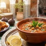 A steaming bowl of tomato-based Moroccan Harira soup with lentils and chickpeas, served with lemon, crusty bread, figs, and Chebakia pastries in a traditional setting.