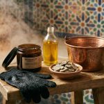 A still life photo in a steamy room showing Moroccan hammam essentials: a jar of black soap, a Kessa exfoliating glove, Ghassoul clay, Argan oil, and a copper bucket on a wooden stool.