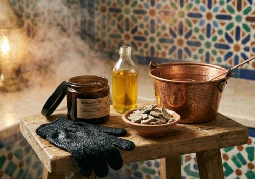 A still life photo in a steamy room showing Moroccan hammam essentials: a jar of black soap, a Kessa exfoliating glove, Ghassoul clay, Argan oil, and a copper bucket on a wooden stool.