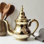 Handcrafted brass teapot in unadorned gold finish styled with wooden spoons on a neutral linen surface