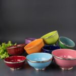 Scattered ceramic bowls with silver trim in bright colors arranged on dark background with lettuce