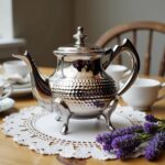 Silver wide oblate hammered teapot with cabriole feet on a white lace doily beside purple flowers