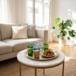 A collection of mixed-color beldi tea glasses arranged on a traditional round brass tray alongside sweet Moroccan pastries, placed on a marble coffee table in an airy living room.