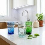 Blue, clear, and green beldi tea glasses featuring decorative silver hardware sitting elegantly on a bright white quartz countertop in a modern, sunlit kitchen.