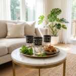 A set of beautiful black pattern tea glasses arranged on a traditional round brass tray with sweet Moroccan pastries and fresh mint, resting on a marble coffee table in a sunlit living room.