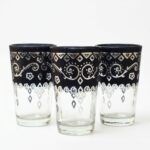 A bright studio close-up of three traditional black pattern tea glasses, showcasing their intricate silver geometric and floral designs against a clean white background.