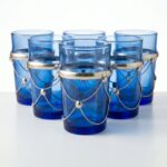 Six vibrant blue beldi tea glasses adorned with intricate silver metal accents and delicate chains, displayed beautifully on a pure white surface.