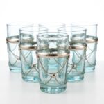 A beautiful set of six clear beldi tea glasses featuring decorative silver metal bands and draped chains, arranged in a triangle against a clean white studio background.