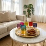 A beautiful set of Colored Glass Tea Cups arranged on a traditional round brass tray with sweet Moroccan Chebakia pastries, resting on a marble coffee table in a sunlit living room.