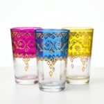 A bright studio close-up of three traditional Moroccan Colored Glass Tea Cups in pink, blue, and yellow, highlighting their beautiful hand-painted gold detailing on a clean white background.
