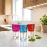 Three vibrant Colored Glass Tea Cups featuring intricate gold painted patterns, elegantly displayed on a bright white quartz kitchen countertop next to fresh mint leaves.