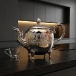 A shiny engraved silver teapot resting on a sleek black marble countertop in a modern, dark-toned kitchen, positioned next to a clear glass teacup.