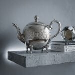 A detailed engraved silver teapot displayed on a modern concrete floating shelf alongside a stack of design books and a geometric glass object against a minimalist grey wall.
