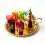 Vibrant gold brass hamsa tea set featuring a traditional engraved teapot with a burgundy tassel, six colorful glasses adorned with gold hamsa hand designs, and a matching round serving tray, isolated on a bright white background.