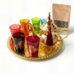 Complete gold brass hamsa tea set beautifully arranged on a serving tray alongside a kraft paper pouch of loose leaf Moroccan green tea on a clean white surface, showcasing the full brewing experience.
