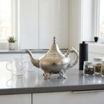 Hammered silver teapot footed traditional placed on a grey countertop with modern glass mugs and tea tins