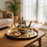 A hamsa engraved silver teapot placed on a decorative round brass tray with almond cookies and Moroccan tea glasses, resting on a wooden coffee table in a bright living room.