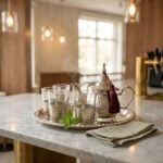 A steaming hand-hammered silver plated tea set displayed on a polished marble countertop in a warm, ambient cafe setting, beautifully accented by a sprig of fresh mint leaves and a folded linen napkin.