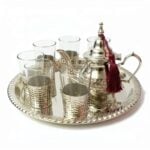 Hand-hammered silver plated tea set featuring an authentic Moroccan teapot adorned with a burgundy tassel, six glasses resting in textured silver bases, and a coordinating round serving tray, isolated on a bright white background.