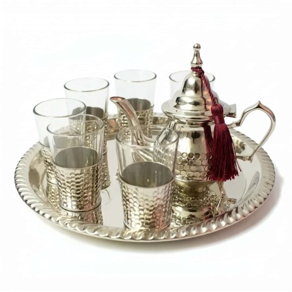 Hand-hammered silver plated tea set featuring an authentic Moroccan teapot adorned with a burgundy tassel, six glasses resting in textured silver bases, and a coordinating round serving tray, isolated on a bright white background.